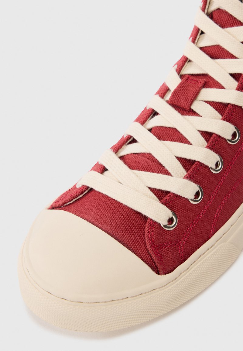 Roter Canvas-Sneaker mit beigem Gummikappen und Sohle, ausgestattet mit beigen Kreuzschnürsenkeln, die durch metalene Ösen gefädelt sind.