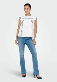 Hvid bluse med blå broderede mønstre, korte cap sleeves, plisseret front, parret med blå udvidede jeans og sorte hæle.
