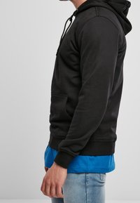 Czarna bluza z kapturem, wykonana z miękkiego materiału, z przednią kieszenią, regulowanym kapturem i ściągaczami; noszona na niebieskiej koszuli.