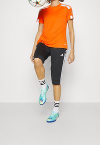 Camiseta atlética de manga corta naranja con acentos blancos, pantalones cortos negros con rayas y logo blancos, zapatos de fútbol en tonos verde azulado y azul con un patrón degradado.
