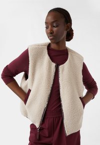 Beżowa kamizelka z polaru o teksturowanym materiale, z otwartym przodem i bocznymi kieszeniami, noszona na bordowej bluzce z długim rękawem.