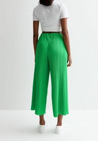 Žena stojící zády, oblečená v bílé zkrácené tričko, jasně zelených skládacích širokých kalhotách a bílých teniskách na jednobarevném pozadí.