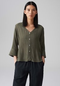 Camicia leggera di colore verde oliva con scollatura a V, chiusura con bottoni e maniche a tre quarti a campana. Tessuto a coste verticali con una vestibilità morbida.