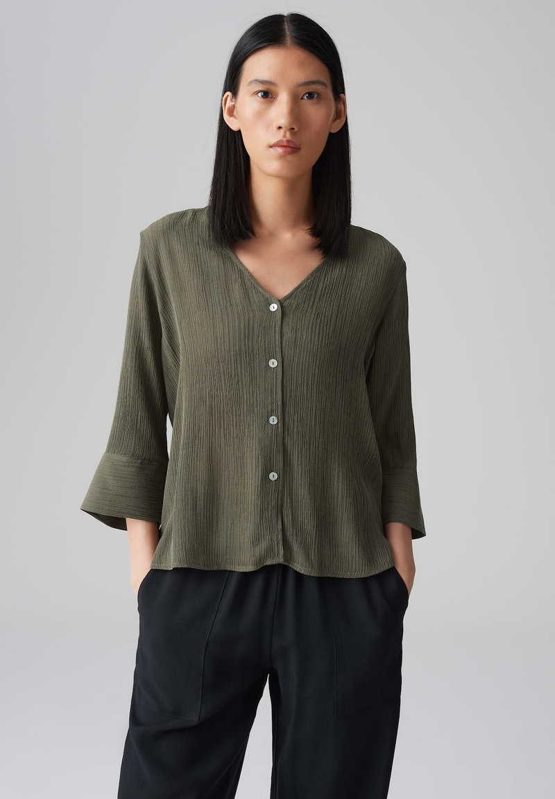 Camicia leggera di colore verde oliva con scollatura a V, chiusura con bottoni e maniche a tre quarti a campana. Tessuto a coste verticali con una vestibilità morbida.