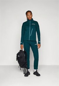 Siniroheline välisjope ja püksid tekstuurse mustriga, helkivad detailid, komplekteeritud musta seljakotiga. Modell seisab ja poseerib.