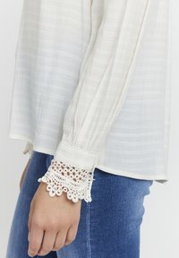 Weiße, strukturierte Bluse mit langen Ärmeln und aufwendiger Spitzenverzierung an den Manschetten, kombiniert mit dunkelblauen Jeans.