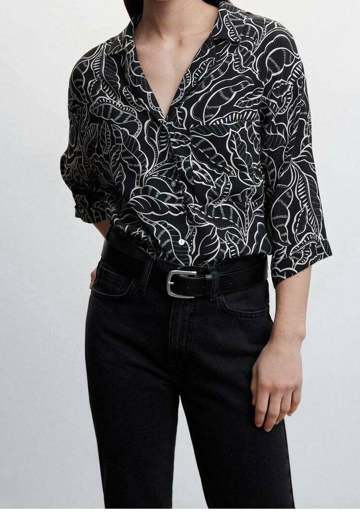 Chemise noire à motif de feuilles blanches, style boutonné, manches longues avec poignets retroussés ; portée avec un jean noir et une ceinture.