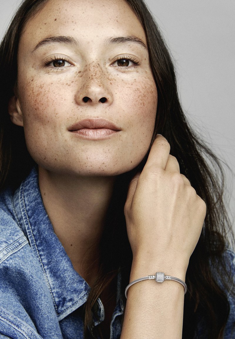 Bracelet en argent avec un fermoir texturé, porté sur une main reposant sur un visage avec une veste en denim. La peau présente des taches de rousseur et une texture lisse.