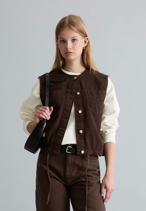 Young woman wearing a white long-sleeve shirt, dark brown buttoned vest, matching pants, holding a black shoulder bag against a gray background.