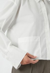 Persona che indossa una camicia bianca a maniche lunghe con ampi polsini e tasca frontale, tiene l'orlo della camicia con una mano sopra pantaloni grigi texture.