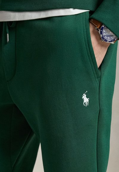 Persoon draagt donkergroene joggingbroek met wit polo speler logo op dij, hand in zak en blauwe horloge met bruine leren band zichtbaar.