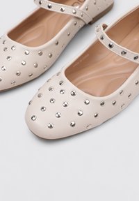 Zapatos de cuero beige con punta redondeada, con tachuelas plateadas redondas y una correa decorativa a lo largo del empeine. Diseño de suela plana.