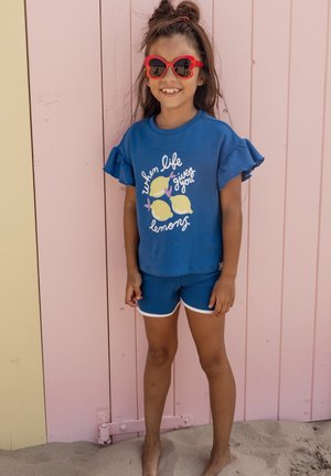 Meisje met rode hartvormige zonnebril lacht terwijl ze een blauw shirt met ruches aan de mouwen en citroenenprint en bijpassende korte broek draagt, staand op zand bij een pastelkleurige muur.