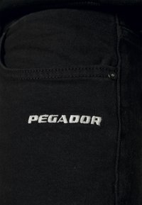 Svart denimtyg har en sydd ficka med texten "PEGADOR" i vit brodyr. Materialet verkar hållbart med en slät yta.