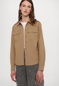 Beige langermet skjorte med knapper, to brystlommer og avrundet kant. Knapper på lommene, kombinert med grå cargo-bukser.