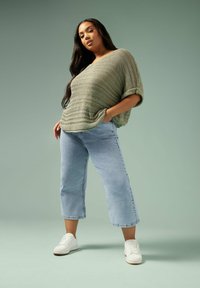 Top suelto de punto en verde oliva con mangas enrolladas, combinado con jeans cortos en azul claro. La modelo lleva zapatillas blancas. Ajuste suave y relajado.