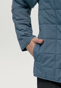 Tejido de chaqueta acolchada azul con una textura suave, que presenta una mano en un bolsillo lateral y un diseño simple con un sutil patrón de cuadrícula.
