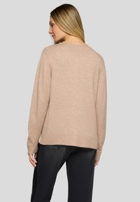 Beiger gestrickter Pullover mit langen Ärmeln und geripptem Saum, der eine lockere Passform und eine weiche Textur zeigt, kombiniert mit dunklen Jeans.