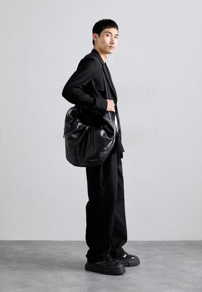 Vyras juodais drabužiais, laikantis didelę juodą odinę rankinę, stovi prieš paprastą šviesiai pilką sieną ir betoninį grindinį.