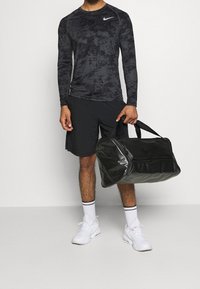 Schwarzes langärmliges Sportshirt mit einem strukturierten Muster, kombiniert mit schwarzen Shorts. Hält eine glänzende schwarze Sporttasche. Weiße Sneakers mit schwarzen Akzenten.