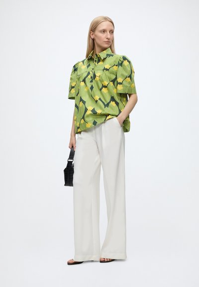 Femme portant un chemisier vert à manches courtes à motif floral, un pantalon blanc à jambes larges, des sandales noires, tenant un petit sac à main noir, debout sur un fond blanc.