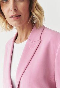 Blazer su misura rosa chiaro con collo a punta e un delicato accento ricamato. Indossato sopra una maglietta bianca, mette in evidenza la texture morbida del tessuto.