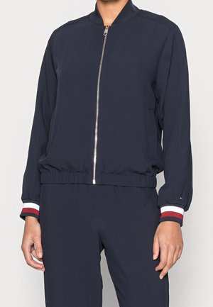 Personne portant une veste bleu marine zippée avec une ceinture et des poignets élastiques ornés d'un liseré rayé rouge, blanc et noir.