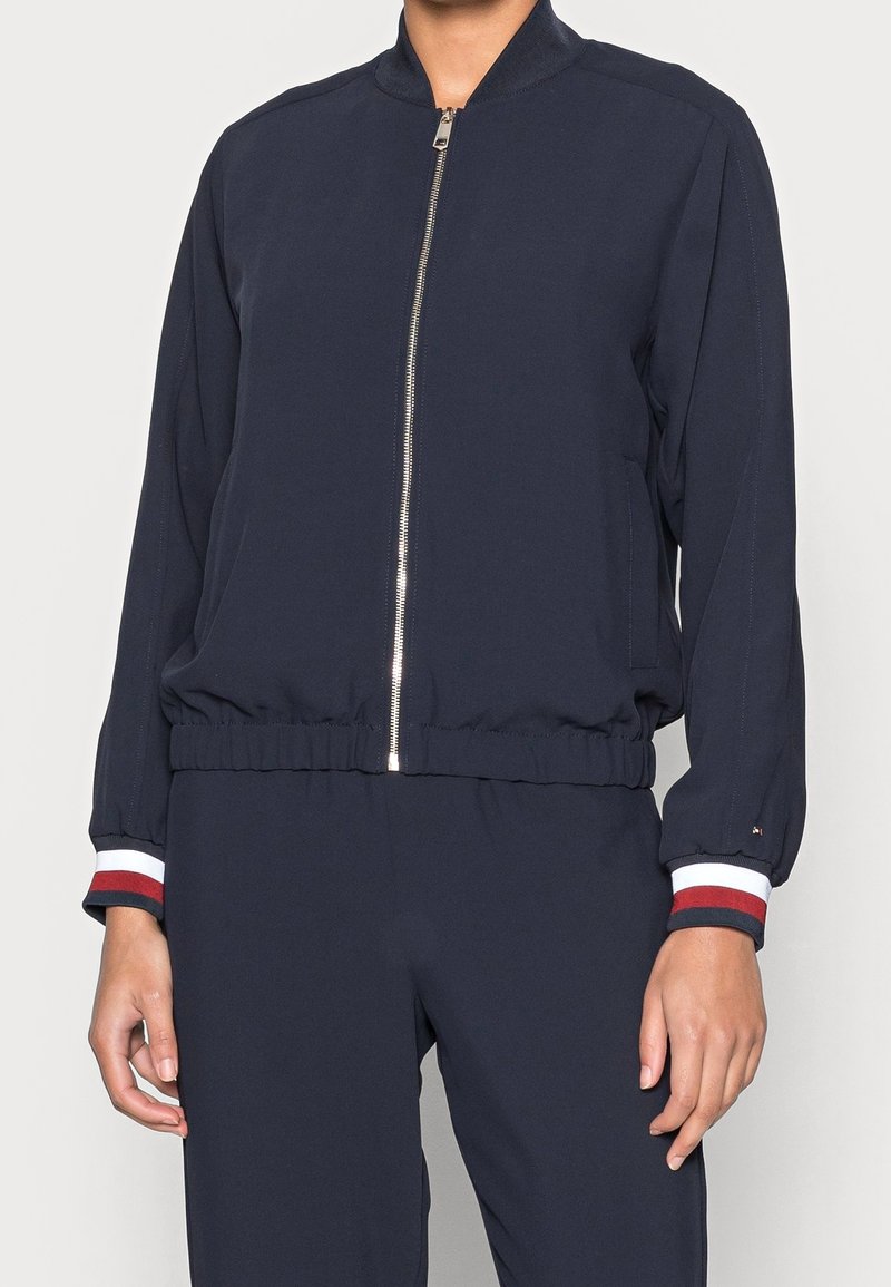 Personne portant une veste bleu marine zippée avec une ceinture et des poignets élastiques ornés d'un liseré rayé rouge, blanc et noir.