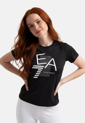 Giovane donna con capelli rossi lunghi che indossa una t-shirt nera Emporio Armani EA7 e pantaloni bianchi, posando con le mani sui fianchi.