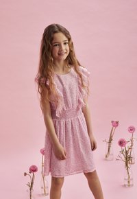 Rosa blommig klänning med volangärmar, smockad midja och knä-lång kjol, mot en rosa bakgrund. Dekorativa vaser med blommor i närheten.