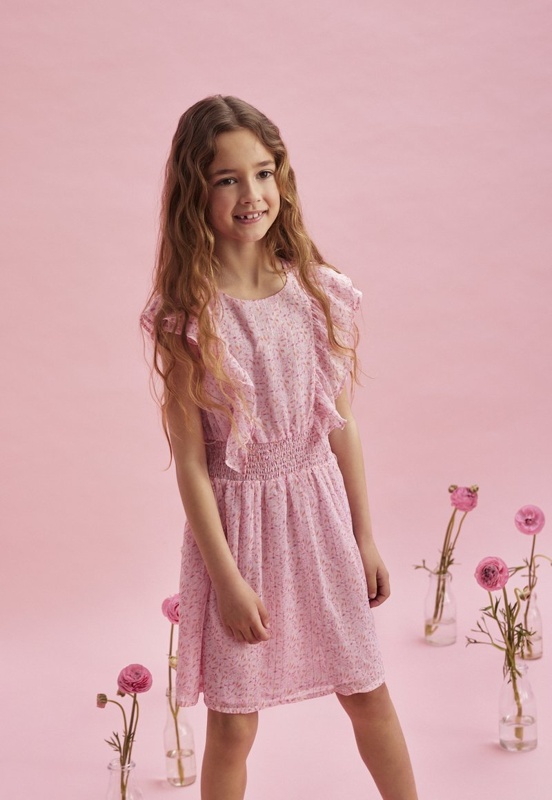 Rosa blommig klänning med volangärmar, smockad midja och knä-lång kjol, mot en rosa bakgrund. Dekorativa vaser med blommor i närheten.