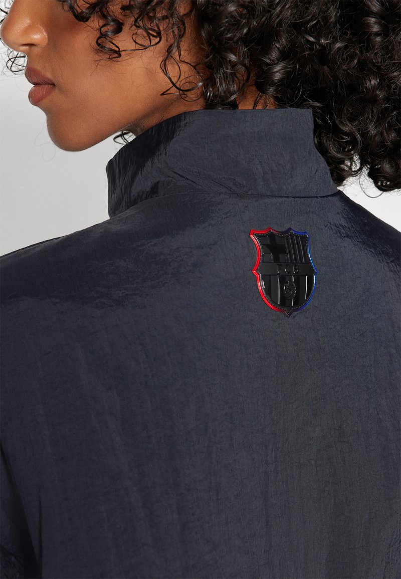 Dunkelblaue, strukturierte Jacke mit hohem Kragen, die auf dem Rücken ein schwarzes, rotes und blaues Wappen-Logo trägt. Sie hat ein glattes, leichtes Aussehen.