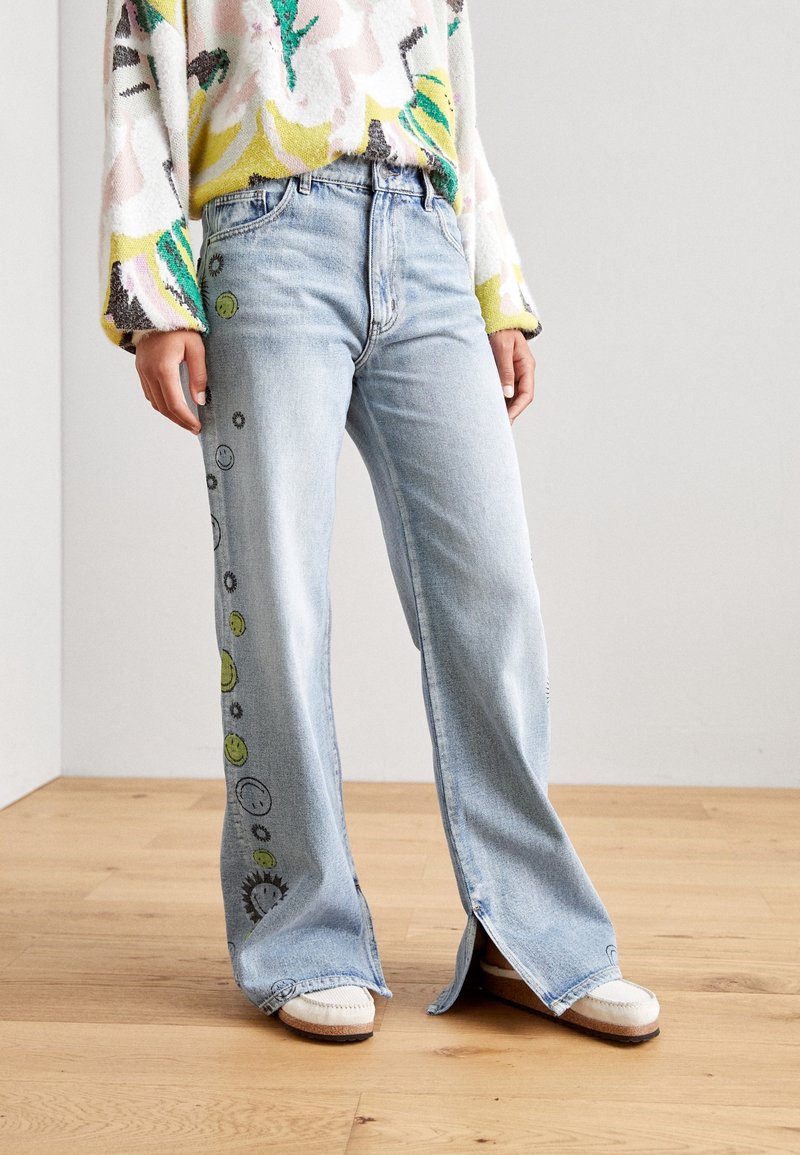 Jeans de pierna ancha en azul claro con caras sonrientes bordadas y diseños de sol a lo largo del costado, con una ligera abertura en el dobladillo.