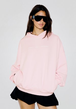 Femme portant de grandes lunettes de soleil noires, un sweat à capuche rose pâle oversize avec les mains dans les poches et une jupe noire courte devant un fond uni.