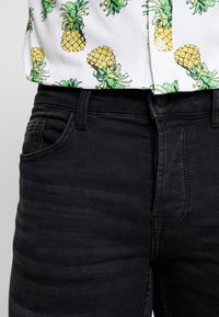 Camisa de manga corta con piñas amarillas y hojas verdes sobre un fondo blanco, combinada con unos vaqueros negros de diseño clásico de cinco bolsillos.