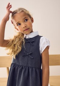 Grå denimkjole med sløjfedetaljer, lagdelt over en hvid skjorte med krave. Kjolen har en skørt med vidde og en tætsiddende liv.