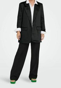 Blazer en satin noir avec revers crantés, deux poches avant, associé à un pantalon noir à jambes larges et des sandales plateformes vertes. Chemise blanche boutonnée visible.