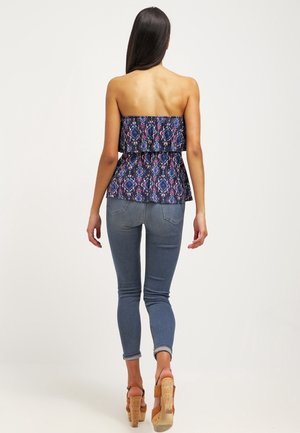 Woman standing facing away, wearing strapless patterned top, fitted blue jeans, and tan high-heeled sandals with cork soles.