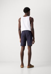 Marineblauwe zwembroek met een gestructureerd ontwerp en een elastische tailleband, gecombineerd met een witte tanktop. Bruine instapschoenen.