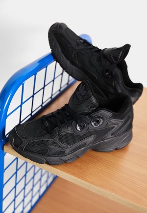 Para czarnych sportowych sneakersów z siateczkowymi i skórzanymi detalami spoczywająca na drewnianej półce z niebieską metalową kratownicą.