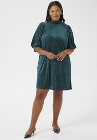 Türkisfarbenes, kurzärmeliges Kleid aus geripptem Samt mit hohem Kragen, lässigem Schnitt und puffärmeligen Ärmeln, kombiniert mit schwarzen spitzen High Heels.