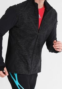 Corredor masculino vestido con una chaqueta negra con cremallera sobre una camiseta roja de Adidas, con mallas negras que presentan patrones geométricos azules.