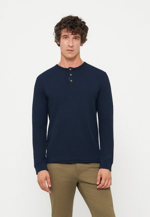 Homme aux cheveux bouclés portant un t-shirt henley bleu marine à manches longues et un pantalon marron clair, debout devant un fond blanc uni.