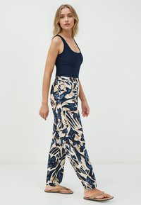 Femme blonde marchant, portant un haut sans manches bleu marine, un pantalon large à motifs beige et bleu, et des sandales plates beige avec une boucle en or.