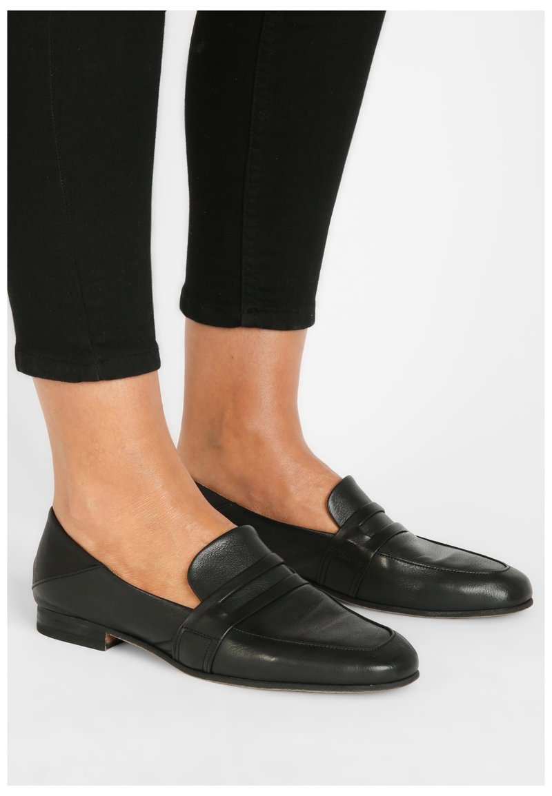 Mocassins en cuir noir avec une semelle plate, présentant un détail de bride sur le dessus ; design élégant et minimaliste avec une texture lisse.