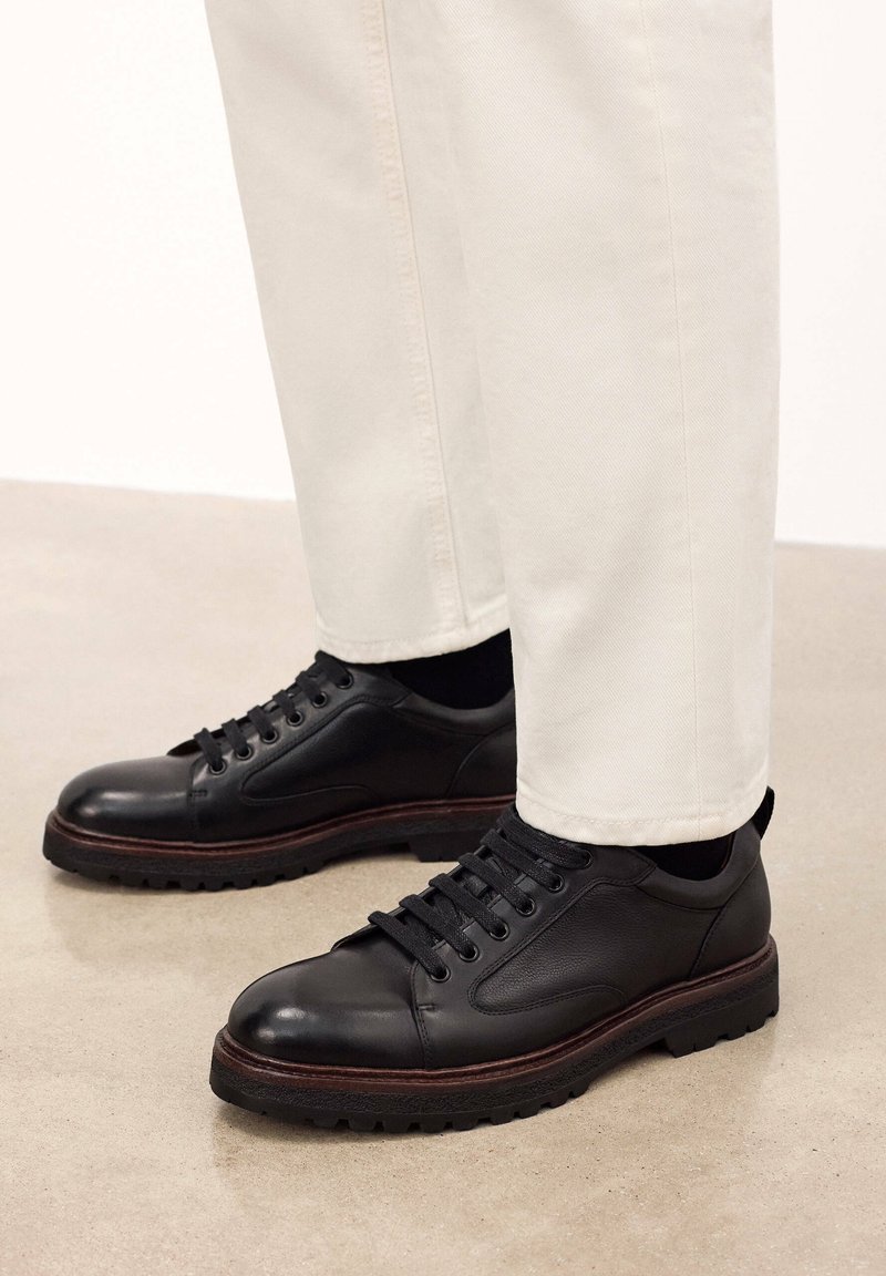 Scarpe in pelle nera con punta arrotondata e superficie testurizzata, dotate di chiusura con lacci e una suola intermedia marrone a contrasto. Indossate con pantaloni di colore chiaro.