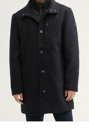 Uomo che indossa un cappotto di lana blu scuro abbottonato con tasca sul petto con cerniera e colletto interno della giacca nera visibile su sfondo chiaro.