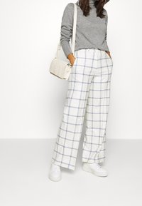 Weite Karottenhose in Weiß mit blauen Karos, kombiniert mit einem grauen Strickpullover und weißen Sneakers; ergänzt mit einer cremefarbenen Umhängetasche.