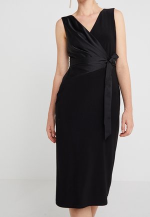 Robe midi noire sans manches avec col en V, détail en satin drapé et ceinture nouée à la taille portée par une personne debout sur un fond uni.