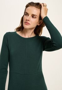 Frau mit rotem Haar, die ein dunkelgrünes Langarmoberteil trägt, sich an ihrem Haar berührt, vor einem neutralen beigen Hintergrund.